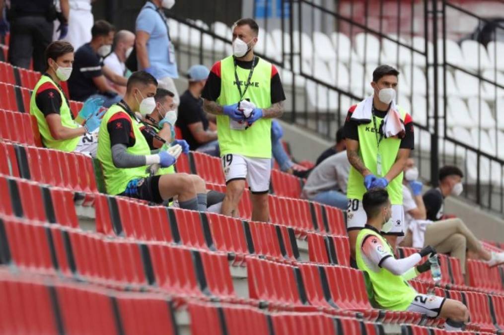 Futbolistas suplentes del Rayo Vallecano, ataviados con guantes y mascarillas, esperan en la grada del Estadio de Vallecas.