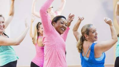 A multi-ethnic group of women having fun taking an exercise class. The instructor is out of focus in the background, wearing a microphone. The main focus is on the African American woman in pink and the Caucasian woman in royal blue, arms raised, laughing in the foreground.