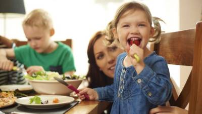 a young family of four sit at the dining table and eat pizza and salad.Mum helps her little girl with her meal , as she cheekily stuffs a whole tomato into her mouth .