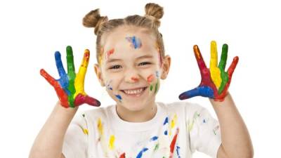 Portrait of a smiling little girl with hands and face covered with different colors of paint. The girl wears a white shirt also spotted with paint and stands against white background. High key DSRL studio photo taken with Canon EOS 5D Mk II and Canon EF 70-200mm f/2.8L IS II USM Telephoto Zoom Lens