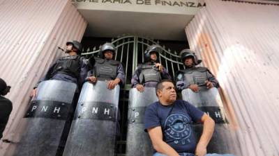 Un maestro está sentado, dando la espalda a la Secretaría de Finanzas que es custodiada por cuatro agentes de la Policía Nacional de Honduras.