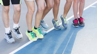 Cropped view of group of teenagers standing on a tennis court wearing sneakers. There are five pairs of legs on the lines of the hardcourt. The shoes are different colors, some with bright shoe laces. They are in a row, moving their feet.