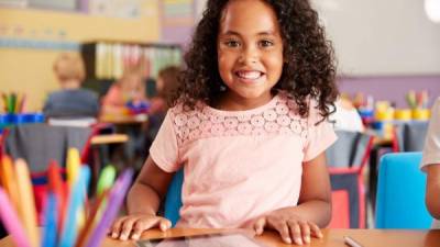 Cheerful Caucasian preschool student in her classroom. She is wearing a backpack and a school uniform. She is holding a book.