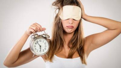 Beautiful young woman with sleeping mask holding alarm clock. She is tired and lazy in morning.