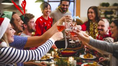 Group of friends toast together with their glasses of wine. They are sitting around a table with food and christmas crackers in from of them. Wearing party hats and novelty jumpers, tinsel ext they celebrate together.