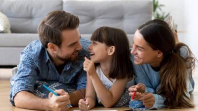 Diverse multi-ethnic young family married couple small adorable daughter lying on wooden warm floor at modern home drawing on paper use colored pencils together have fun spend free time on weekend