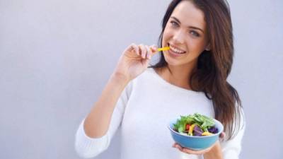 Portrait of an attractive young woman standing against a gray background and eating a bowl of salad