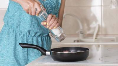 Woman grinding salt from a salt mill into a frying pan as she cooks the dinner, close up of her hands and the pan