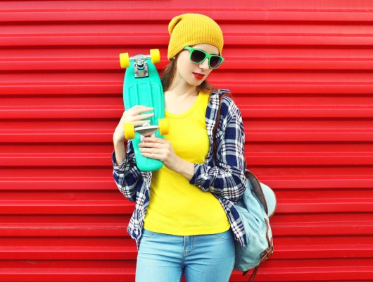 Fashion portrait of stylish girl in sunglasses and colorful clothes with skateboard over red background