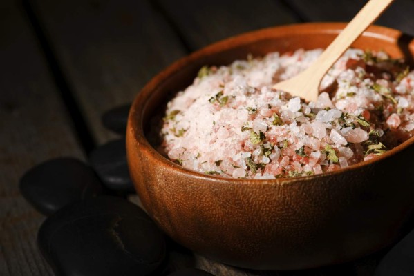 Horizontal photo of wooden bowl full of bath pink sea salt with herbs inside. Bowl is placed on old wooden board with few black stones around.