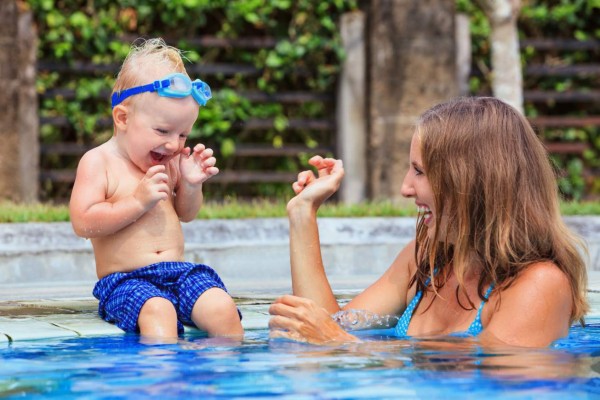 Little swimming child in underwater goggles sit on poolside, has fun - baby splashing with mother in pool. Family lifestyle and summer holiday water sports outdoor activity and lessons with parents.