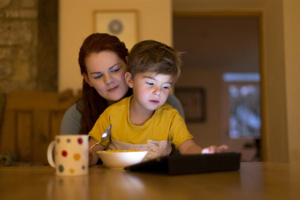 Woman sits with a little boy on her knee at a table. The boy is using a digital tablet while eating cereal and the woman watching closely over his shoulder.