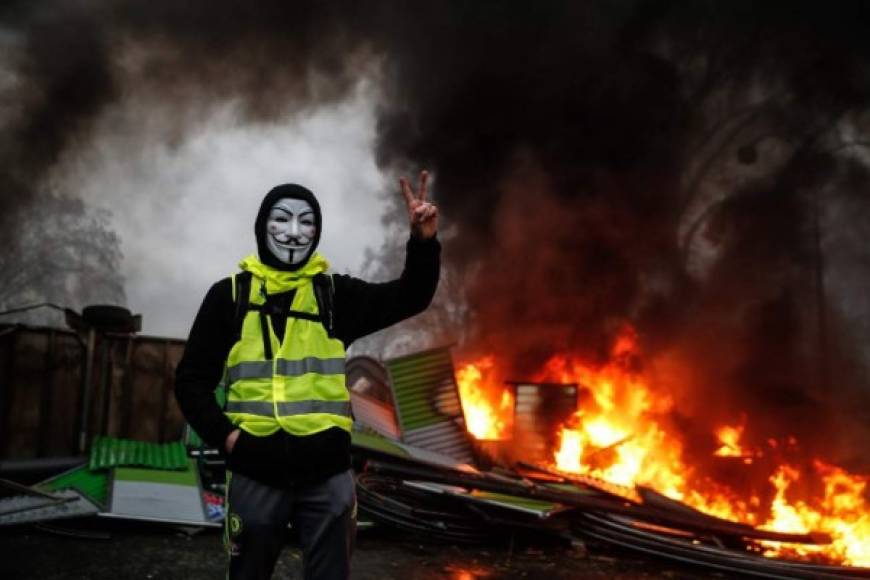 A protester wearing a Guy Fawkes mask makes the victory sign near a burning barricade during a protest of Yellow vests (Gilets jaunes) against rising oil prices and living costs, on December 1, 2018 in Paris. (Photo by Abdulmonam EASSA / AFP)