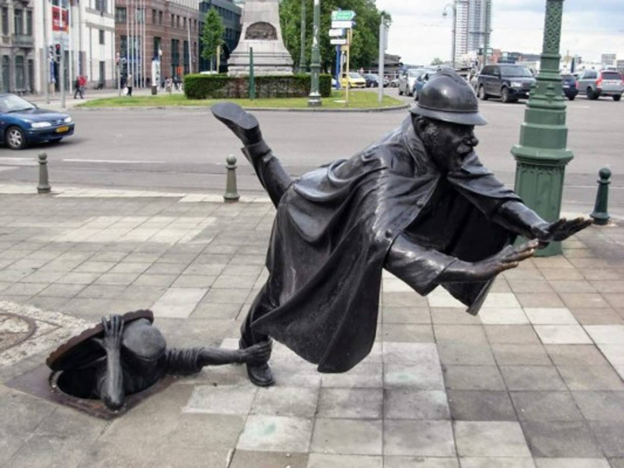 'De Vaartkapoen' se halla en Bélgica. La obra exhibe a dos hombres: uno está en una alcantarilla y toma al otro del pie, quien al parecer es un agente de policía. <br/>Imagen tomada de http://detallelogia.blogspot.com/