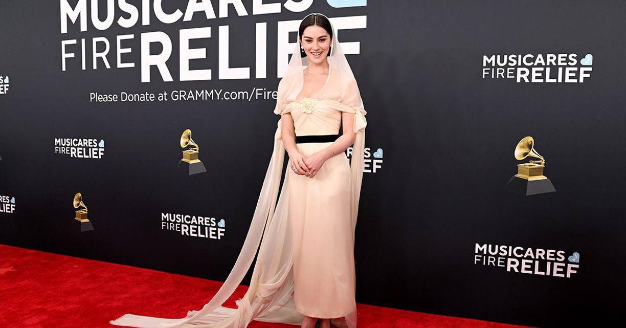 <b>Gracie Abrams</b> llevó un vestido largo en tono marfil con un diseño fluido y minimalista. La prenda incluía un cinturón negro que marcaba la cintura y aportó contraste al conjunto.Complementó el look con un velo traslúcido que cubría su cabello y caía suavemente sobre los hombros, evocando una estética romántica y etérea. Completó el estilismo con zapatos negros de tacón bajo y un peinado recogido que realzó la simplicidad elegante del atuendo.