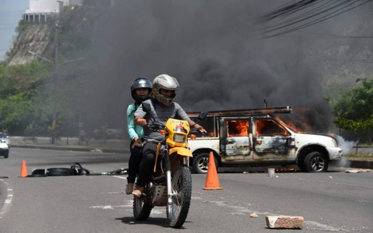 Un hombre conduce una motocicleta y pasa por carro quemado supuestamente por manifestantes en Tegucigalpa.AFP