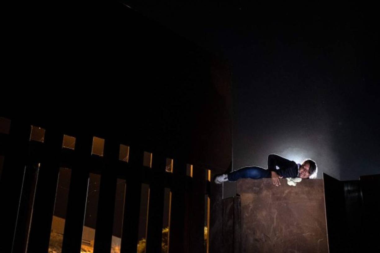 Los migrantes se apoyan en rocas de la desértica barranca y se impulsan unos a otros para trepar la reja de metal oxidado.