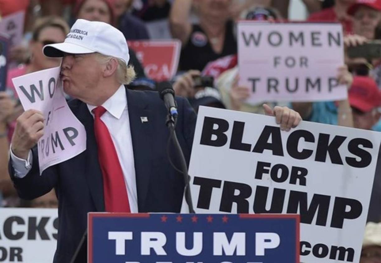 El presidente electo de EUA, Donald Trump, besa un cartel que dice 'mujeres para Trump' y sostiene otro donde se lee: Afroamericanos para Trump, durante un evento de su campaña presidencial en noviembre pasado.