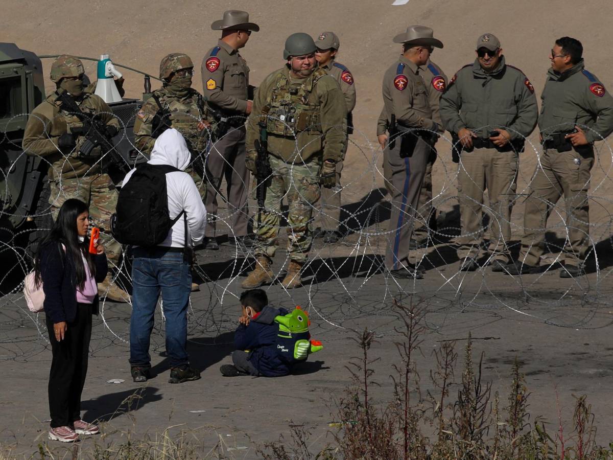 Una familia migrante espera poder cruzar la frontera frente a un grupo de soldados y agentes fronterizos.