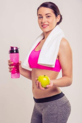 Attractive young woman in sportswear with a towel on her neck is smiling, holding a bottle of water and an apple, on a gray background
