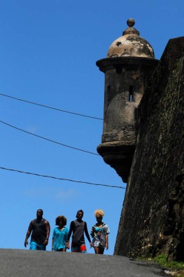 A group of people walk by a 17th Century Spanish Fortress stone walls between El Morro and the fort San Cristobal in the neighbourhood of La Perla where the video 'Suavecito' was recorded in San Juan, on July 22, 2017.Something unusual is happening in La Perla, a poor barrio clinging to a steep hillside between Old San Juan and the sea where the video for the pop hit 'Despacito' was filmed. / AFP PHOTO / Ricardo ARDUENGO