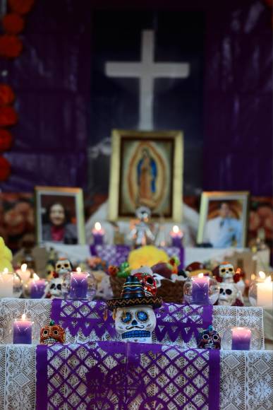 La cruz y la imagen de la Virgen de Guadalupe al fondo posan junto a las fotografías de los difuntos honrados. En un plano más cercano se aprecian las calaveras, un símbolo de la mortalidad y la muerte, que representan a los seres queridos que han fallecido.