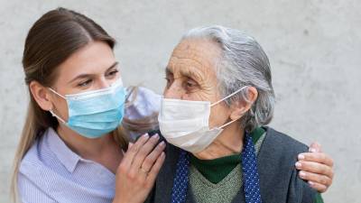 Portrait of friendly caregiver posing with elderly ill woman wearing surgical mask because of covid-19 pandemic