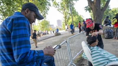 Fotografía que muestra un inmigrante mientras le corta el cabello a otro afuera del refugio de Randall Island en Nueva York.
