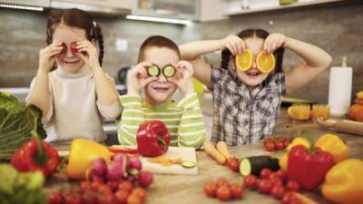 Three little children holding slices of fruit and vegetables over their eyes.