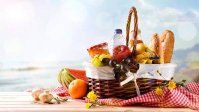 Picnic wicker basket with food on wood table on the beach with blue sky background and sun. Picnic concept. Front view. Horizontal composition.