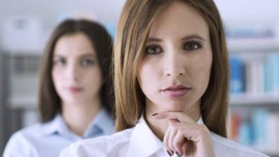 Attractive confident businesswoman posing with hand on chin and looking at camera, her collegue is standing on background