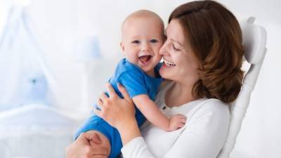 Young mother holding her newborn child. Mom nursing baby. Woman and new born boy in white bedroom with rocking chair and blue crib. Nursery interior. Mother playing with laughing kid. Family at home