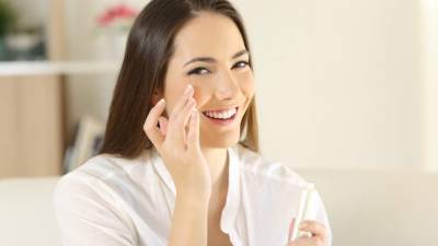 Woman applying moisturizer cream on the face and looking at camera sitting on a couch in the living room at home