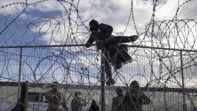 Un migrante escala la cerca alambrada para ingresar a EEUU frente a varios agentes de la Guardia Nacional de Texas.
