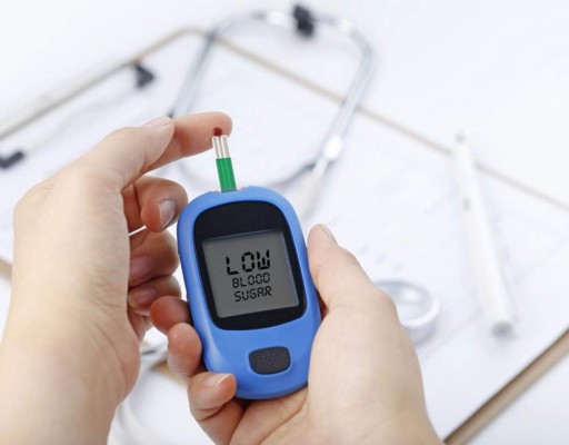 Hand holding a blood glucose meter measuring blood sugar, the background is a stethoscope and chart file