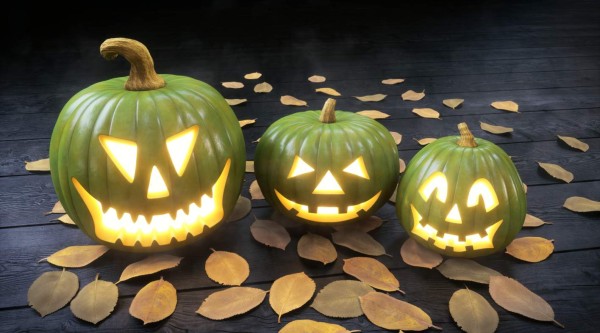 A set of three green colored Jack O' lanterns sitting on a dark wooden floor, surrounded with fallen leaves.