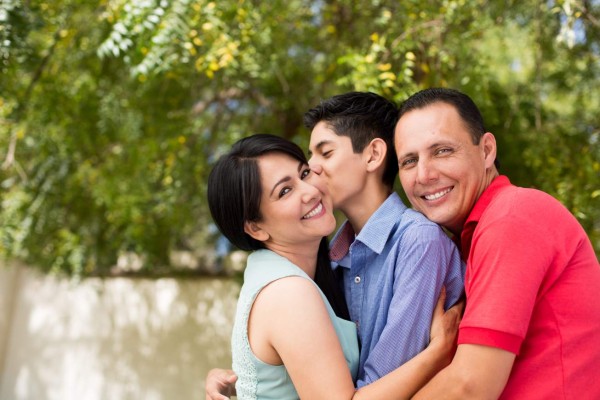 A horizontal shot outdoors of a latin teenage boy between his parents embracing him and smiling at the camera.