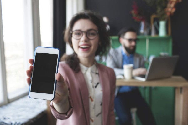 Young woman in glasses showing smartphone and young man working on background