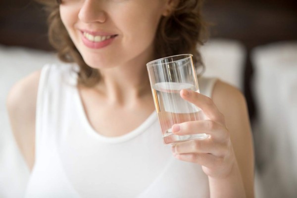 Happy young woman sitting on bed drinking water after getting up. Smiling caucasian female model holding transparent glass in her hand. Close-up. Focus on the arm