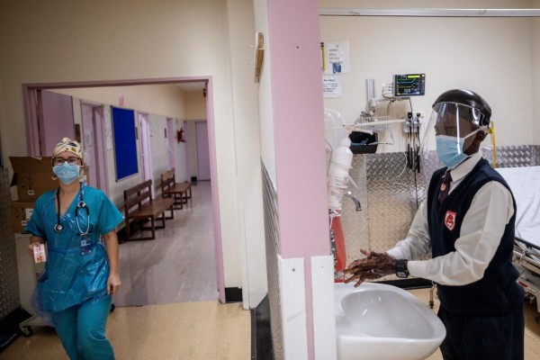 Doctor Jana du Plessis (L) prepares her overall as an health worker washes his hands at the Charlotte Maxeke Hospital in Johannesburg, on April 15, 2020. - A team of medical professionals from the South African public hospital Charlotte Maxeke, lead by Professor Feroza Motara, designed a piece of medical equipment that prevents viral droplets spread to the treating clinicians ensuring safety during intubation, extubation or aerolising procedure. (Photo by Michele Spatari / AFP)