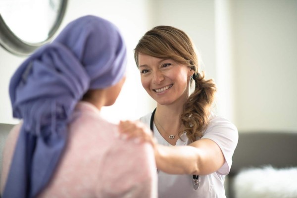 A young woman is indoors in a hospital. She has cancer, and her head is covered with a scarf. She is sitting next to her female doctor. Her female doctor is trying to discuss outcomes with her patient while comforting her.