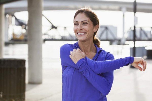 Mature woman taking a break from running to stretch her arms, looking at the view on the waterfront.