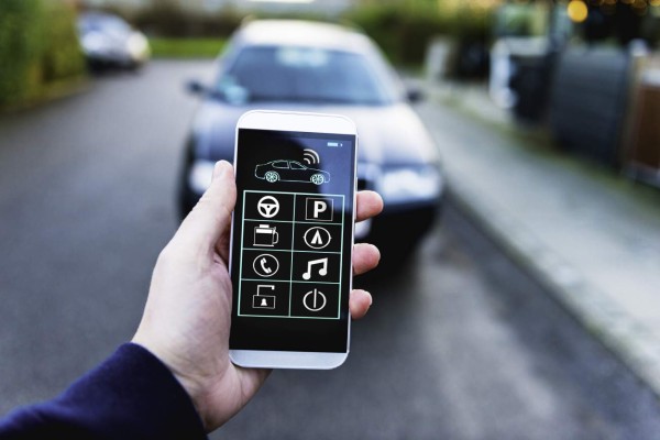 Hand holds a mobile phone with an app that lets the user control different functions of a smart car like parking, playing music, locking and unlocking the vehicle. The phone is connected wirelessly.