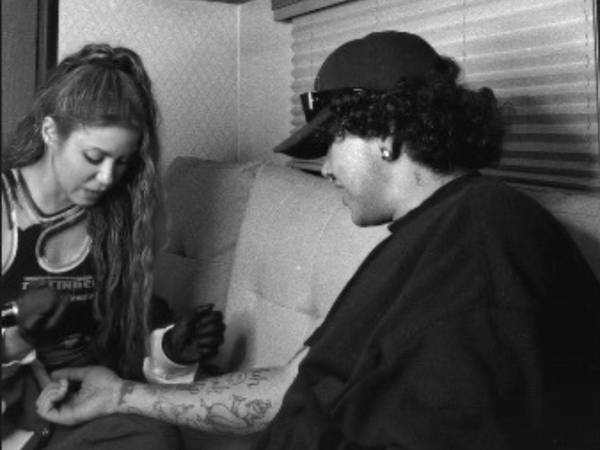 Shakira aparece con guantes negros y una máquina para tatuar mientras toma tinta y dibuja sobre la piel del artista.