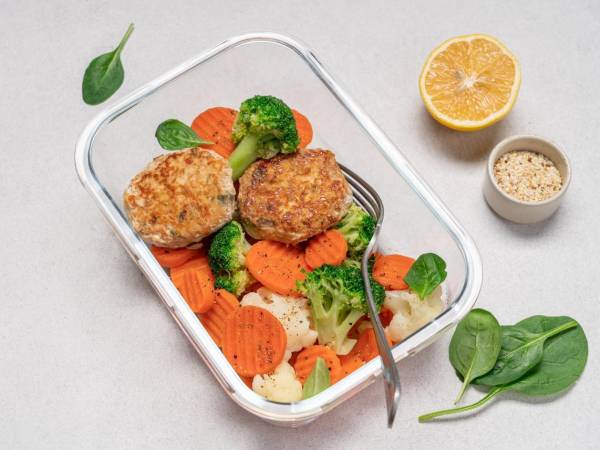 Chuletas de brócoli, zanahoria, coliflor y pechuga de pollo. Comida sana en el envase de cristal en el fondo blanco. Imagen ilustrativa.