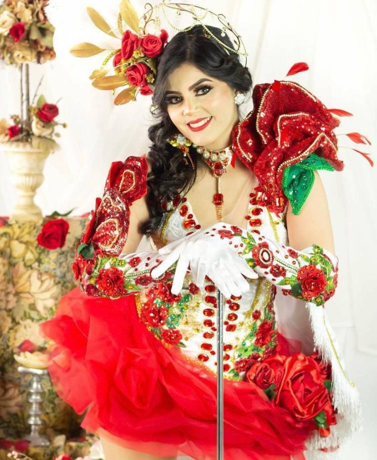 Este hermoso traje lleva en su corset apliques de rosas rojas con pedrería, y como puntos de interés, grandes y llamativos pétalos rojos en ambos hombros, enmarcando el rostro de la bella palillona.