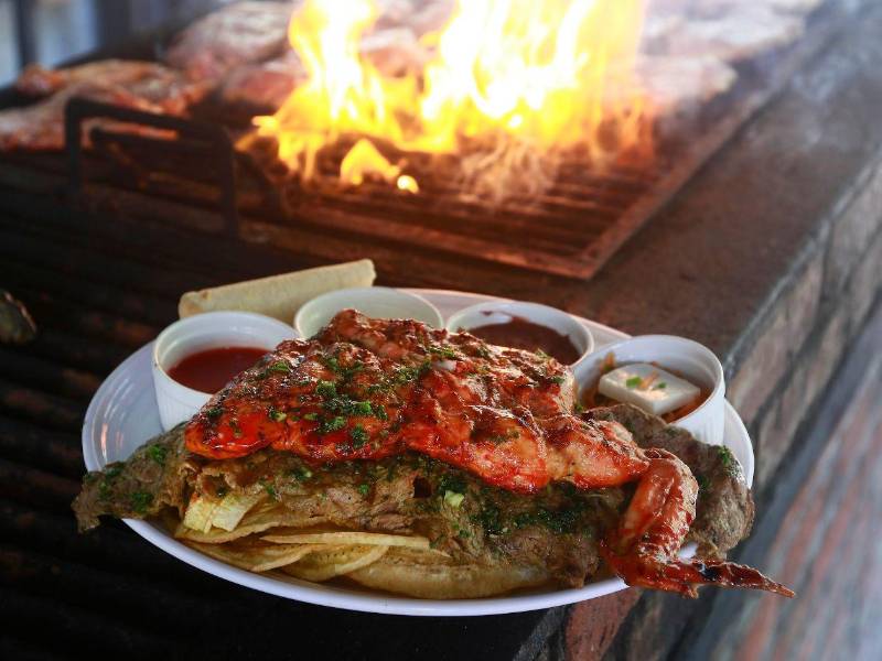 Parrilla encendida y un plato mixto de pollo y carne, acompañado de tajadas, frijoles, queso, encurtido y sus respectivas salsas.