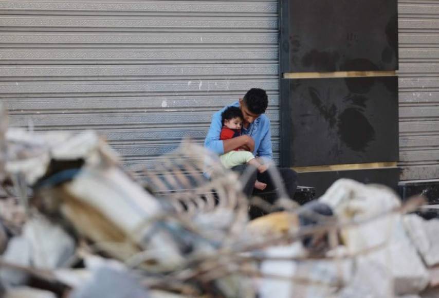 Un palestino sostiene a su hijo mientras está sentado afuera de una tienda cerrada frente a su casa destruida en un vecindario residencial en la ciudad de Gaza a principios del 16 de mayo de 2021, luego del bombardeo masivo israelí del enclave controlado por Hamas. (Foto de MOHAMMED ABED / AFP)