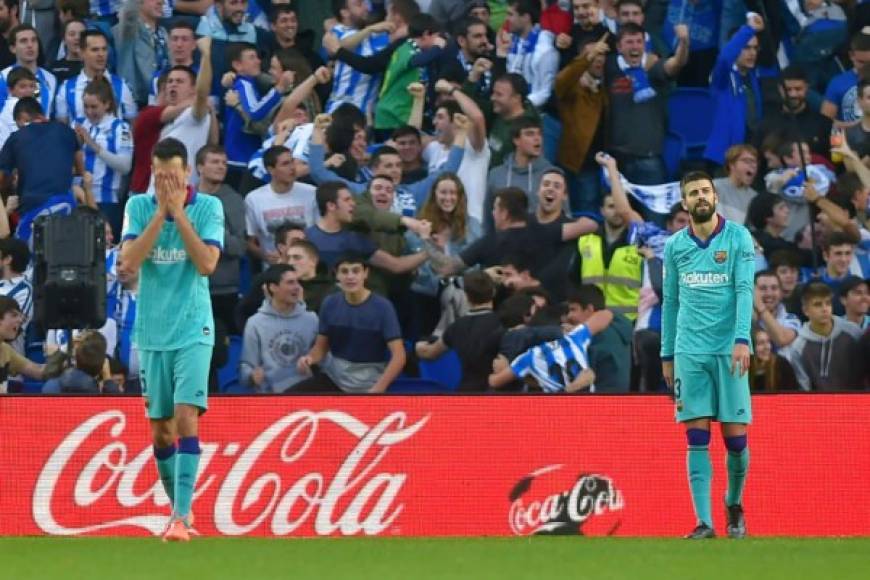 Sergio Busquets se lleva las manos a las cara y Piqué con mirada desencajada luego del primer gol de la Real Sociedad. Al fondo celebran los aficionados locales.