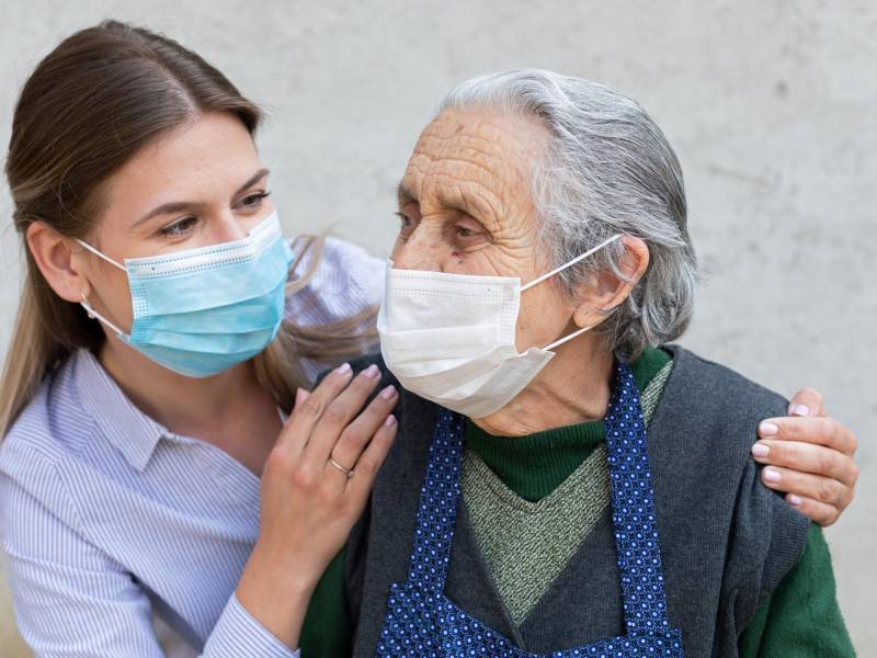 Portrait of friendly caregiver posing with elderly ill woman wearing surgical mask because of covid-19 pandemic